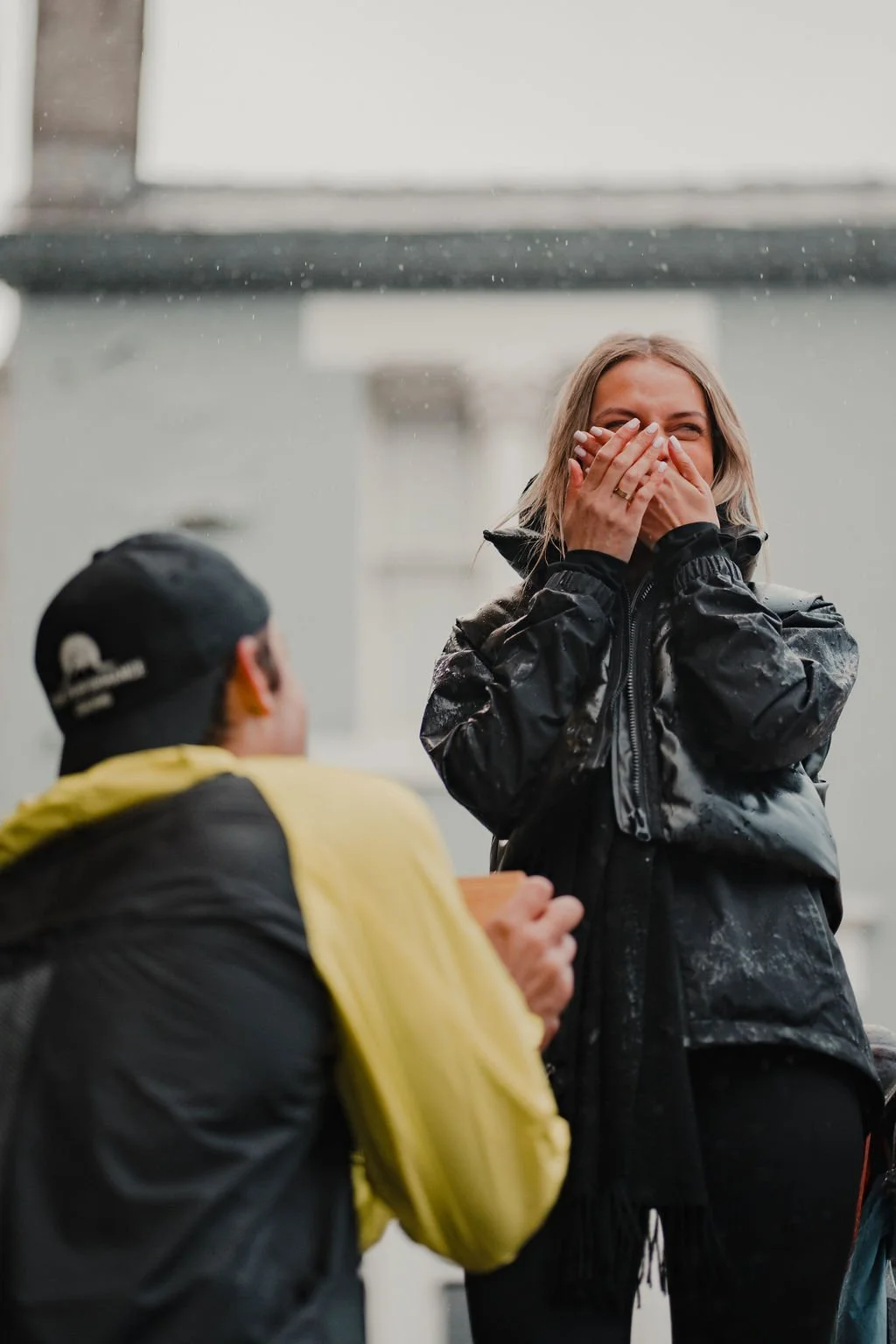A woman with blonde hair laughing and covering her mouth with her hands, wearing a black rain jacket, standing outdoors. A person with dark hair, wearing a yellow and black jacket, is kneeling and facing her.