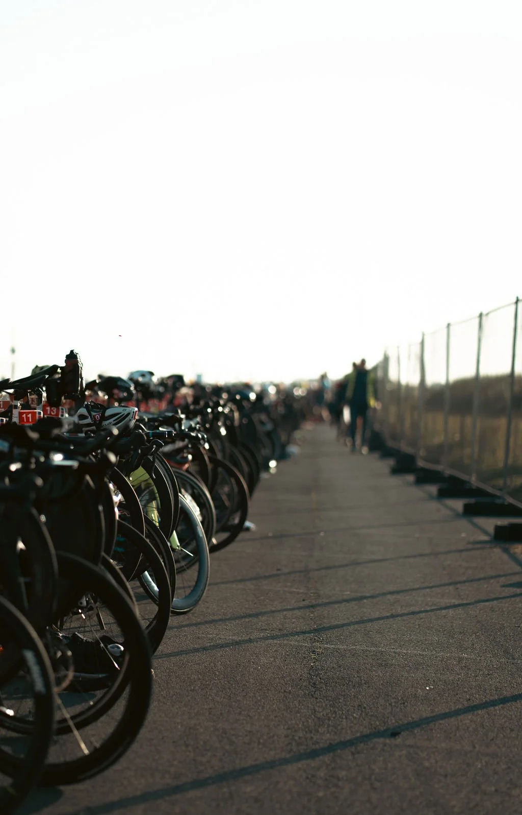 Row of parked bicycles along a paved pathway with a fence on the right side, and a person walking in the distance, outdoors during daylight.