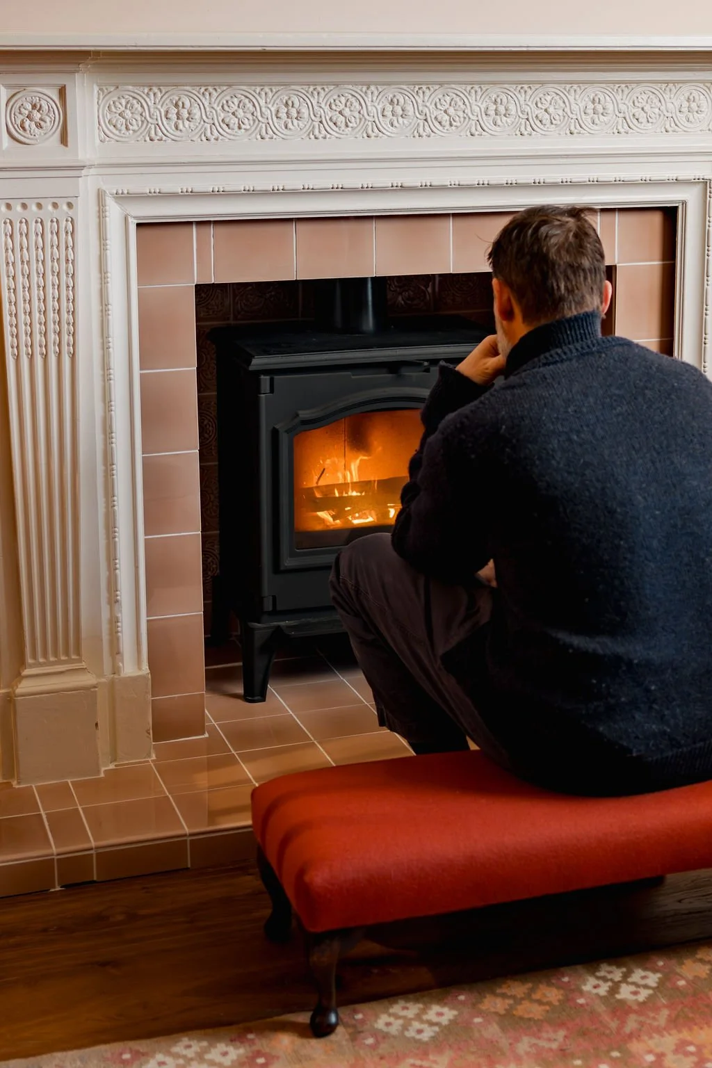 A man is sitting on a small red upholstered stool near a wood-burning stove, warming himself by the fire inside a room with ornate white trim and brown tile accents.