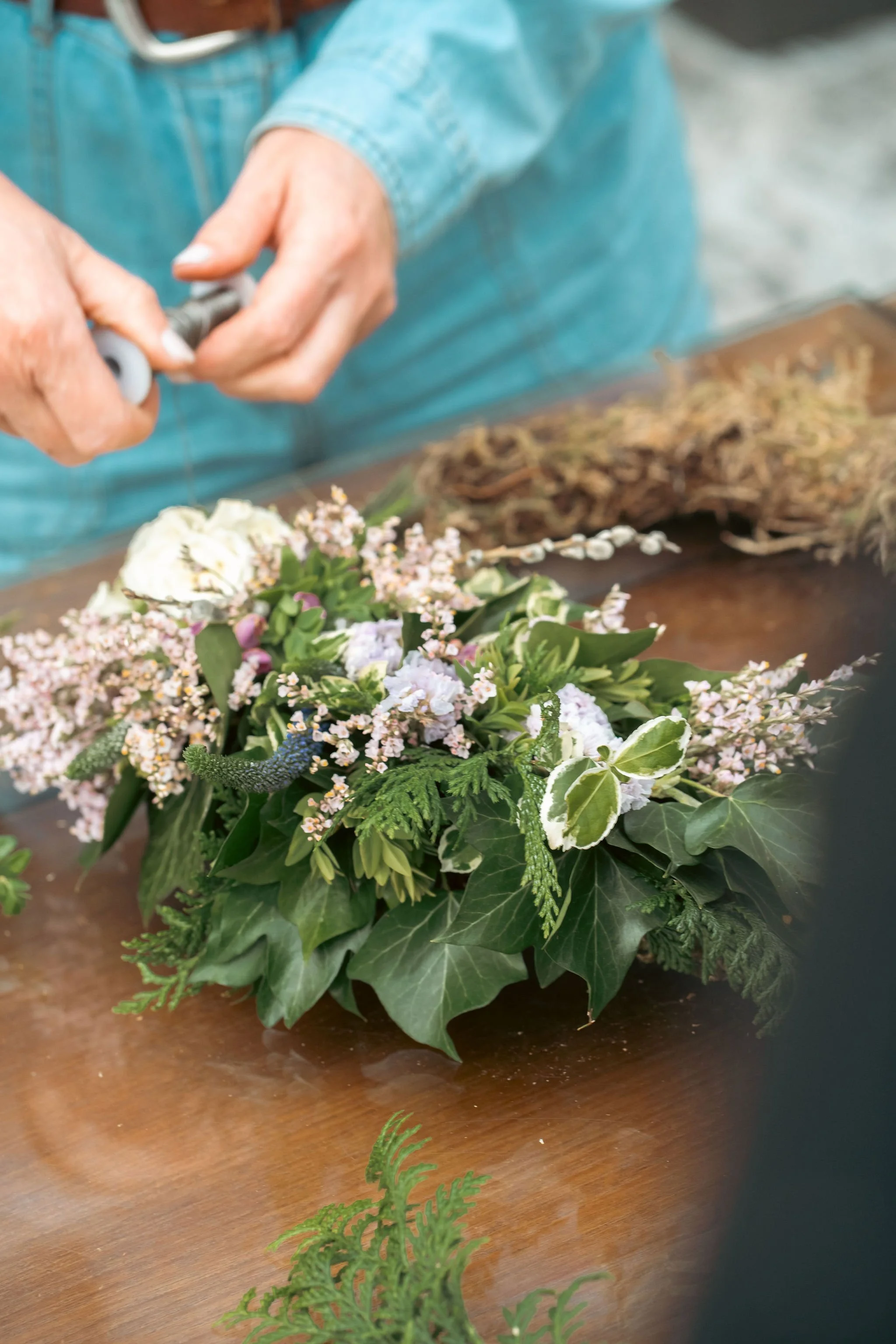 A person decorating a floral arrangement with pink, white, and green flowers and leaves on a wooden table.