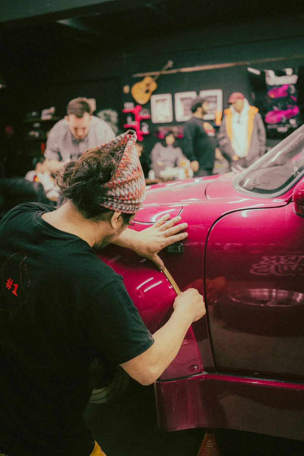 Akira Nakai doing bodywork on RWB build. In the background, there are other people in a workshop or garage, some working and others observing. The workshop has musical instruments, including a guitar hanging on the wall.