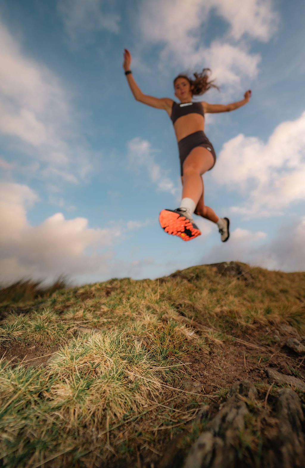 A woman in athletic attire jumping mid-air on a grassy hillside with a cloudy sky background.