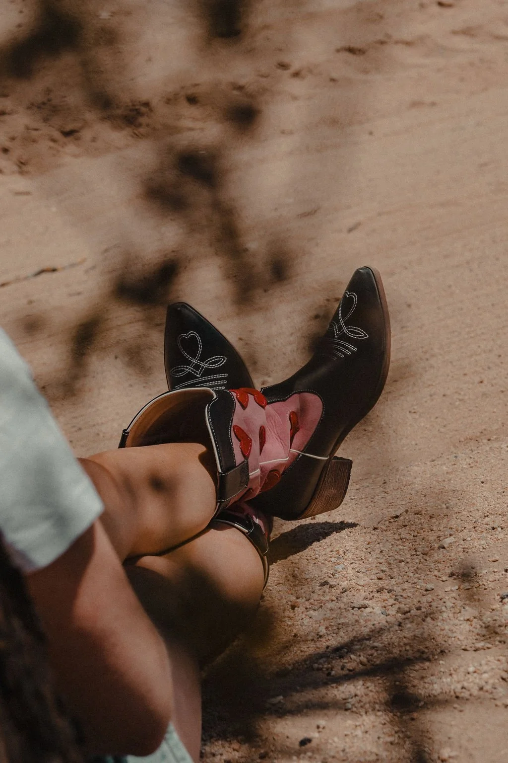 Close-up of a person sitting on sandy ground, wearing black cowboy boots with white embroidery and pink and red accents, and brown heels.