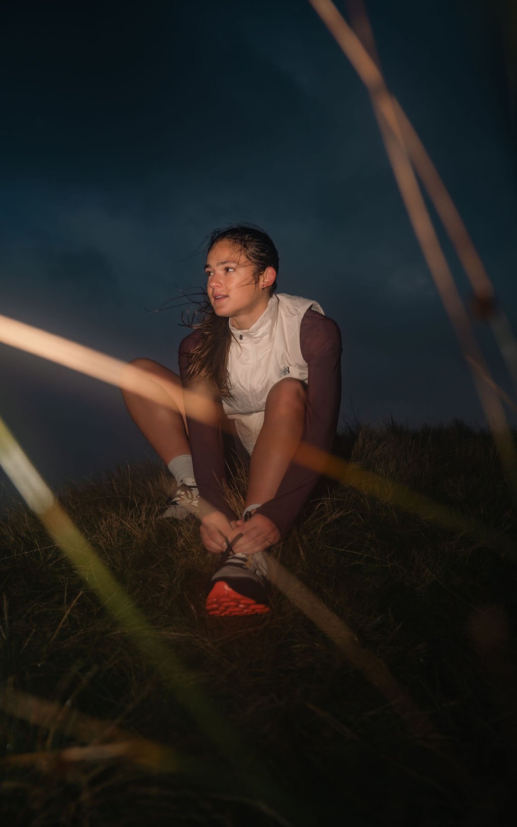 A young woman in athletic clothing sits on grass during dusk, tying her shoelaces with wind-blown hair and a dark cloudy sky overhead.