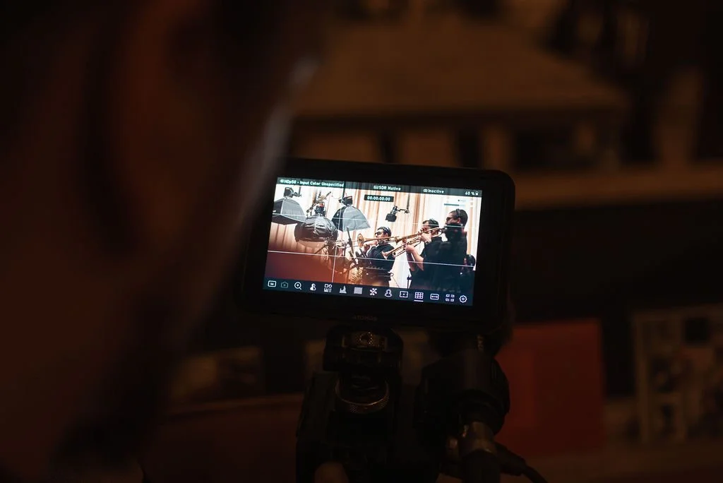 Person filming a live indoor performance of a jazz band playing trumpet and trombone, with a camera viewfinder showing the scene.