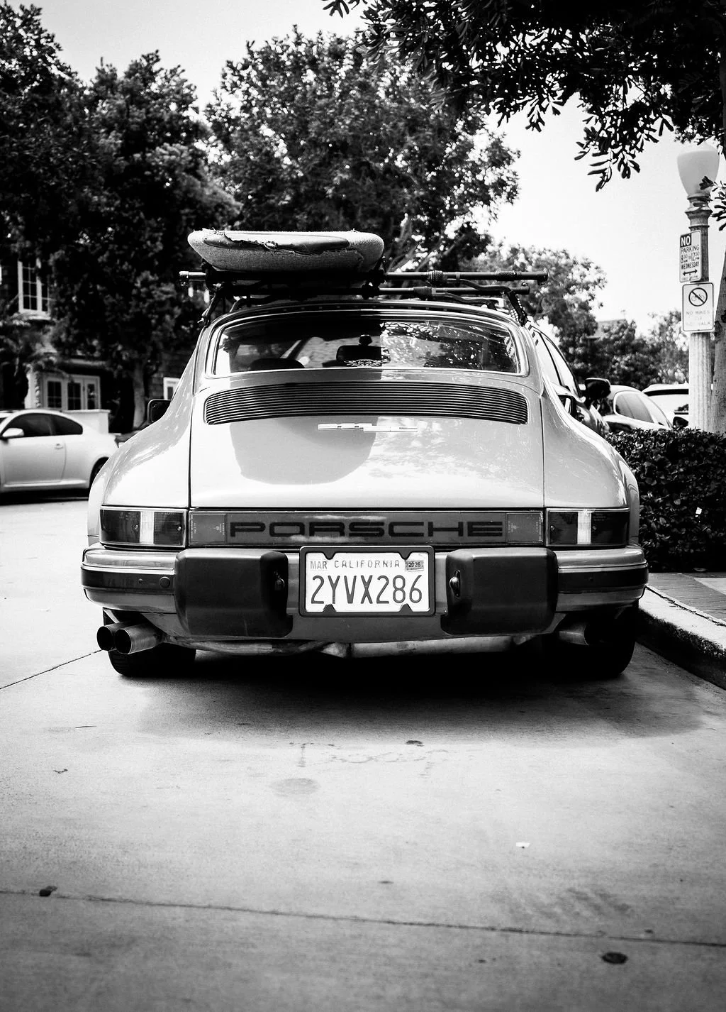 Black-and-white photo of a classic Porsche car parked on a street with a surfboard on top. Surrounding cars, trees, and street signs are visible.