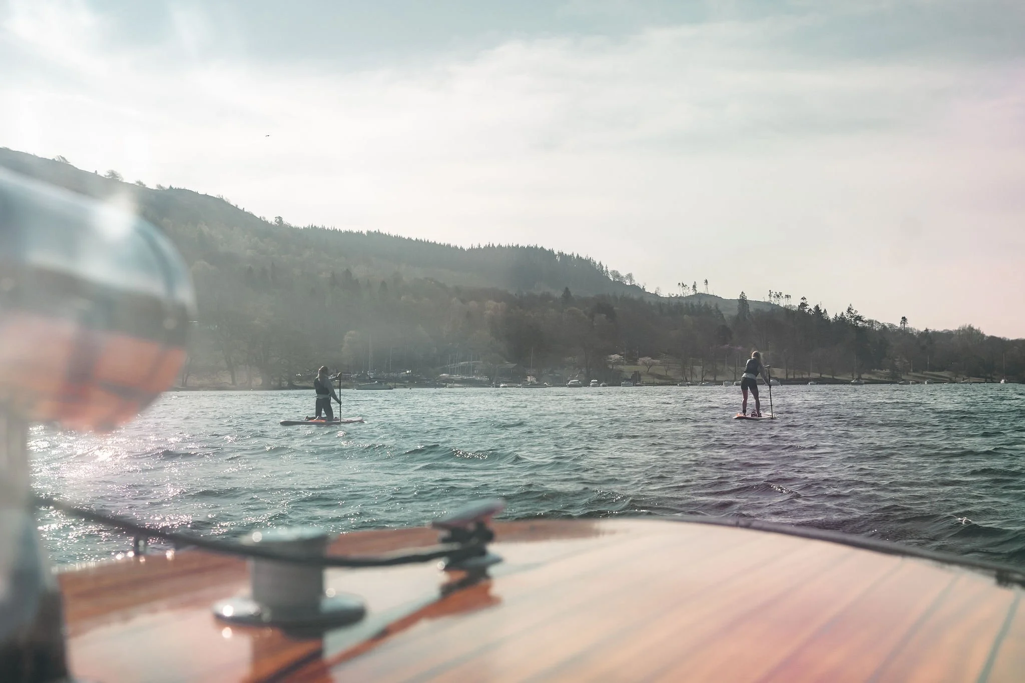 View from a boat on a lake with two people paddleboarding, mountains and trees in the background, and a partly cloudy sky.
