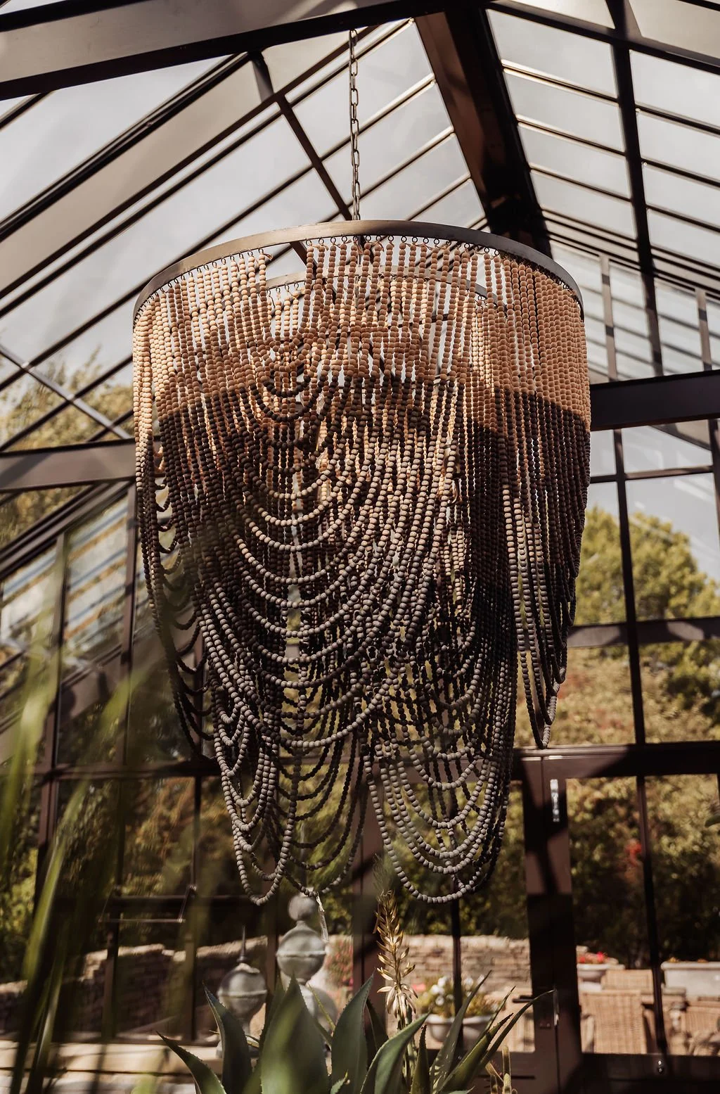 A hanging chandelier made of numerous small beads, with a reflective surface, indoors with large glass windows and greenery outside.