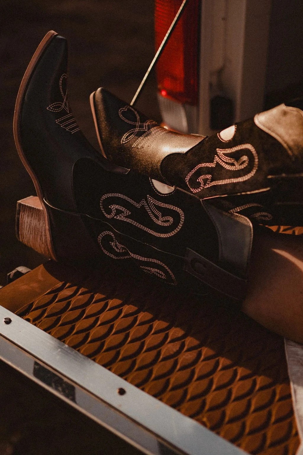 A person is lying on the bed of a pickup truck, wearing cowboy boots with intricate embroidery and a dark outfit. 