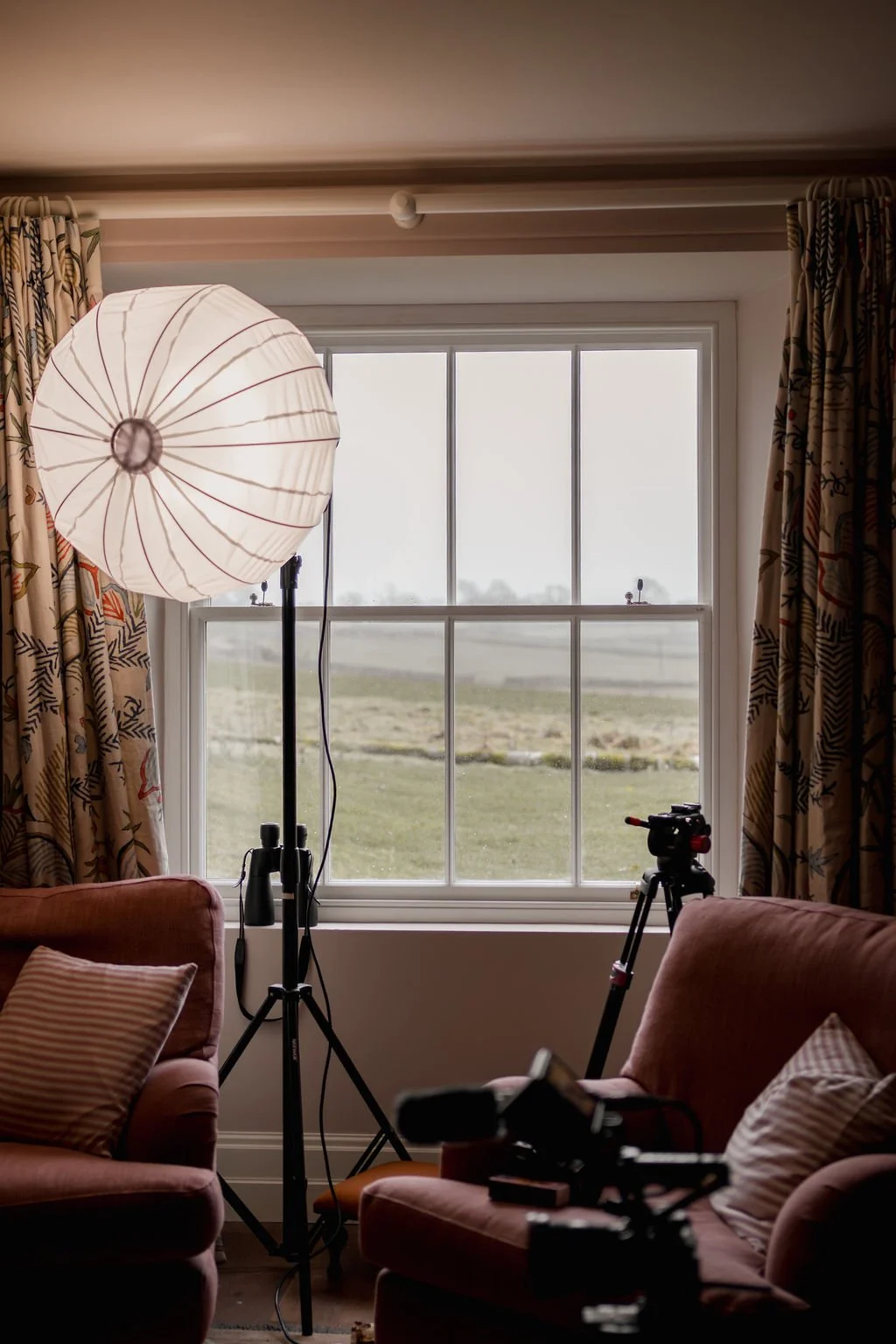 A cozy living room with two pink armchairs, striped cushions, a large window with patterned curtains, a photography studio light, and a camera on a tripod facing the window.