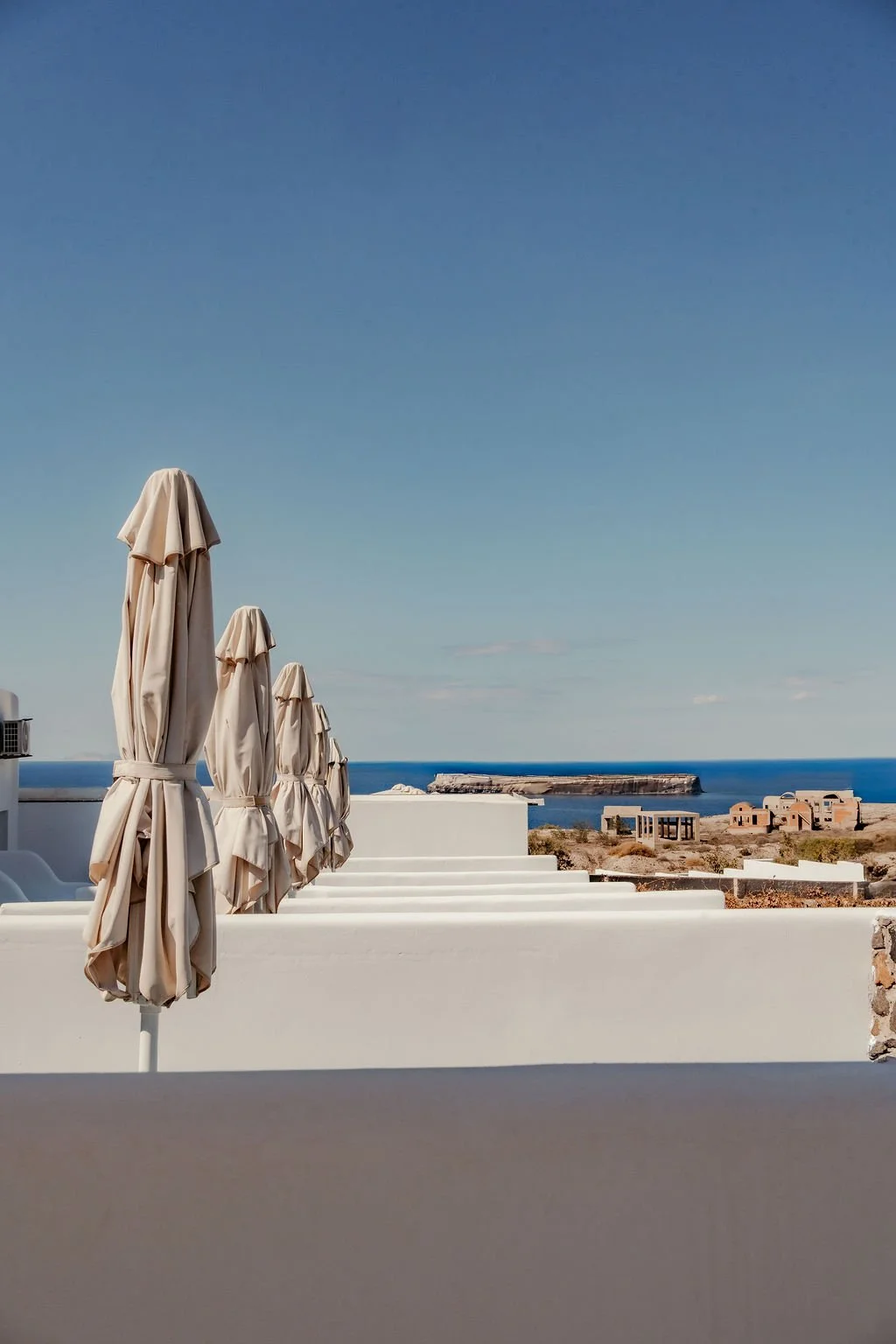 Row of beige patio umbrellas on a white terrace overlooking the sea, with a small island and buildings in the background under a clear blue sky.