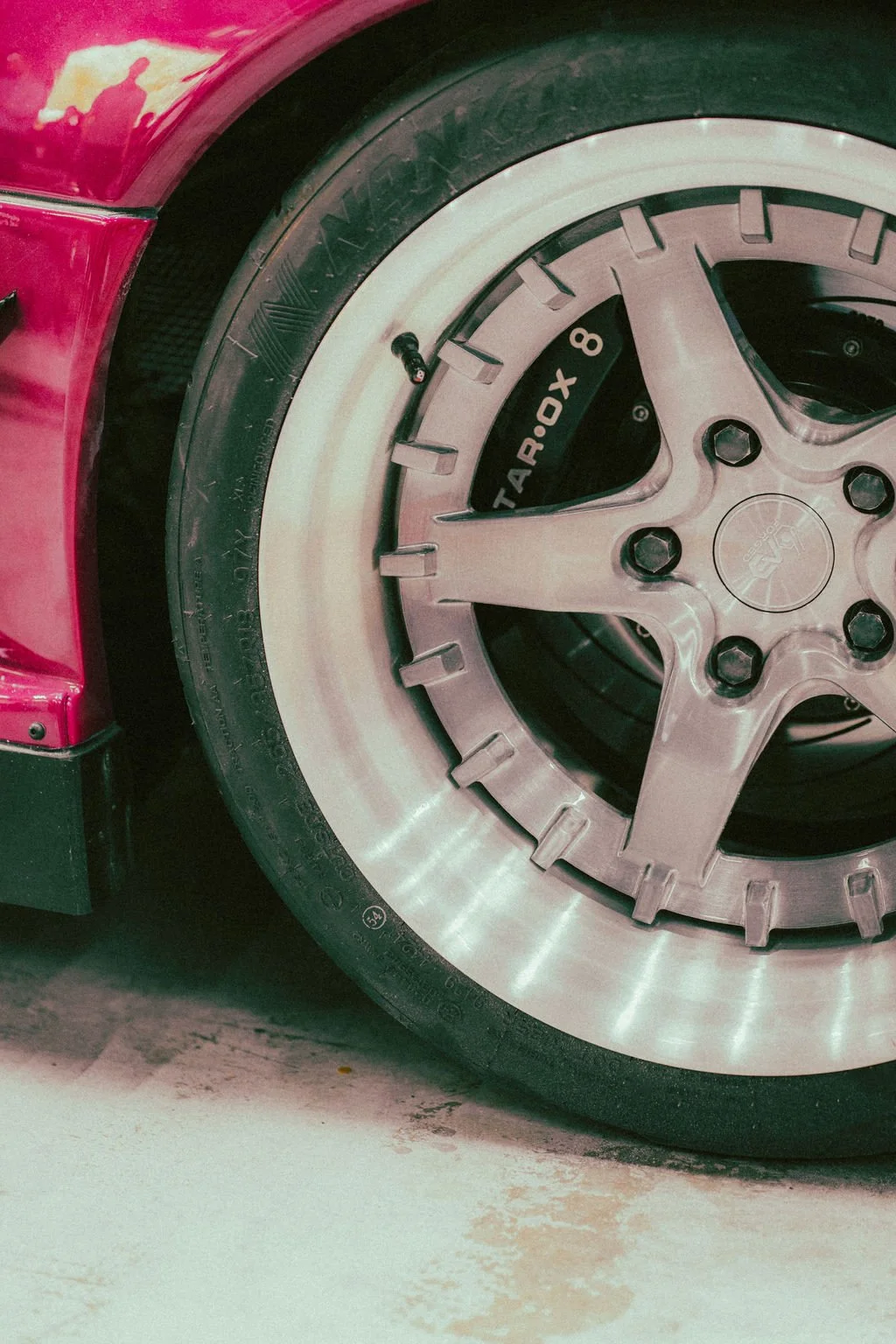 Close-up of a car's wheel, showing a silver rim with a five-spoke design and a black tire with the words 'TAROX' on the brake caliper visible through the rim.