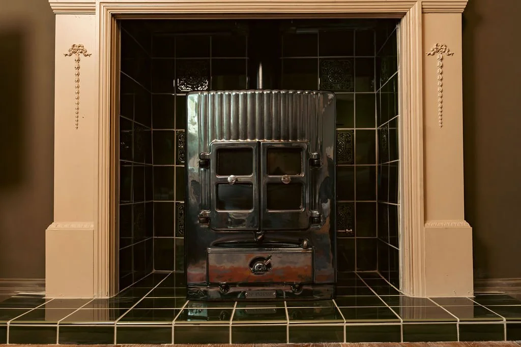 A vintage metal stove with a reflective surface, set into a black-tiled fireplace surrounded by a beige mantel.