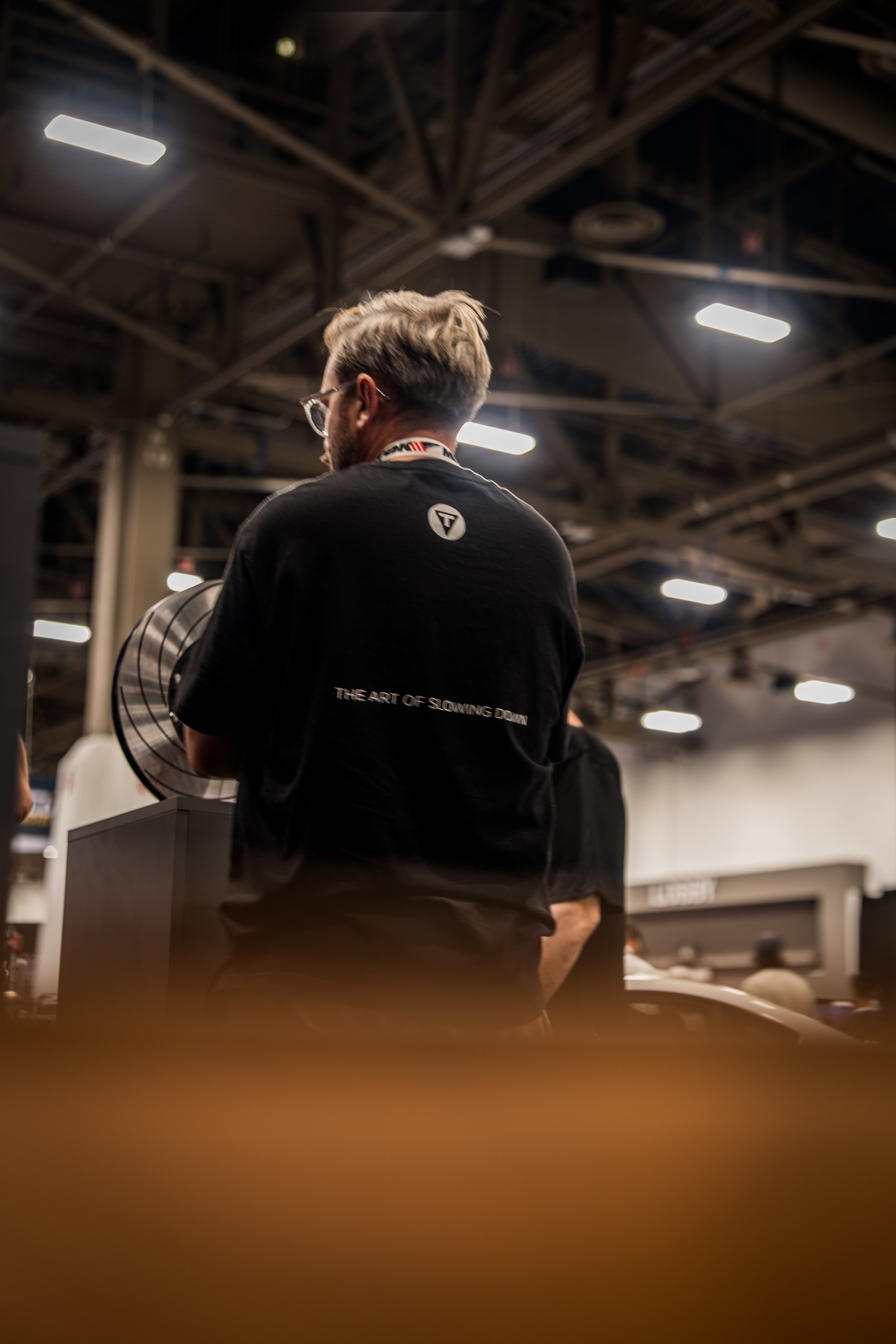 A man with glasses and blonde hair in a black T-shirt, standing indoors in a large, industrial-style space with exposed beams and bright ceiling lights.
