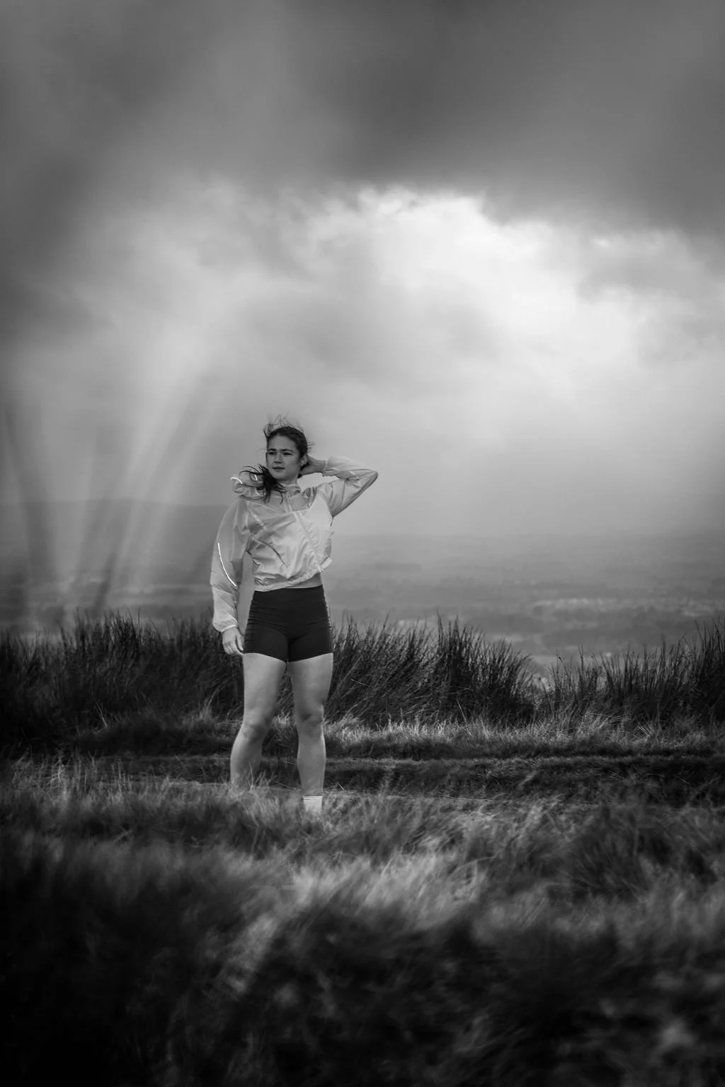 A woman standing on a grassy hilltop under a stormy sky, with one hand behind her head, wearing athletic shorts and a windbreaker, in black and white.