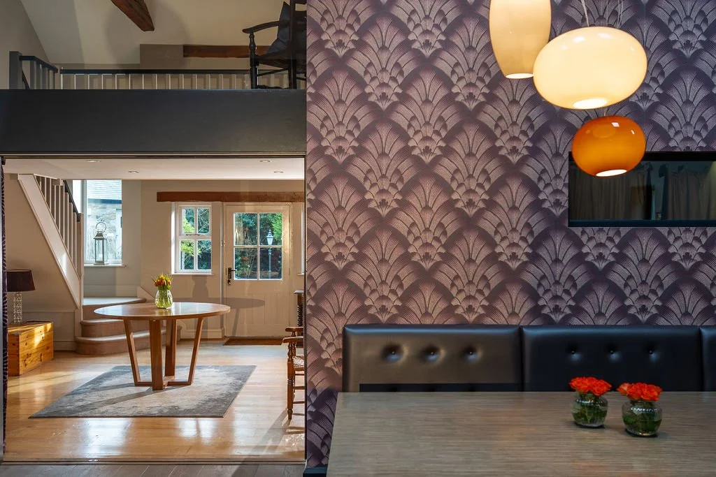 Interior of a modern home with a cozy dining area featuring a leather padded bench, purple patterned wallpaper, and colorful hanging lights, with a view into a bright living space with a round wooden table, windows, and a door leading outside.