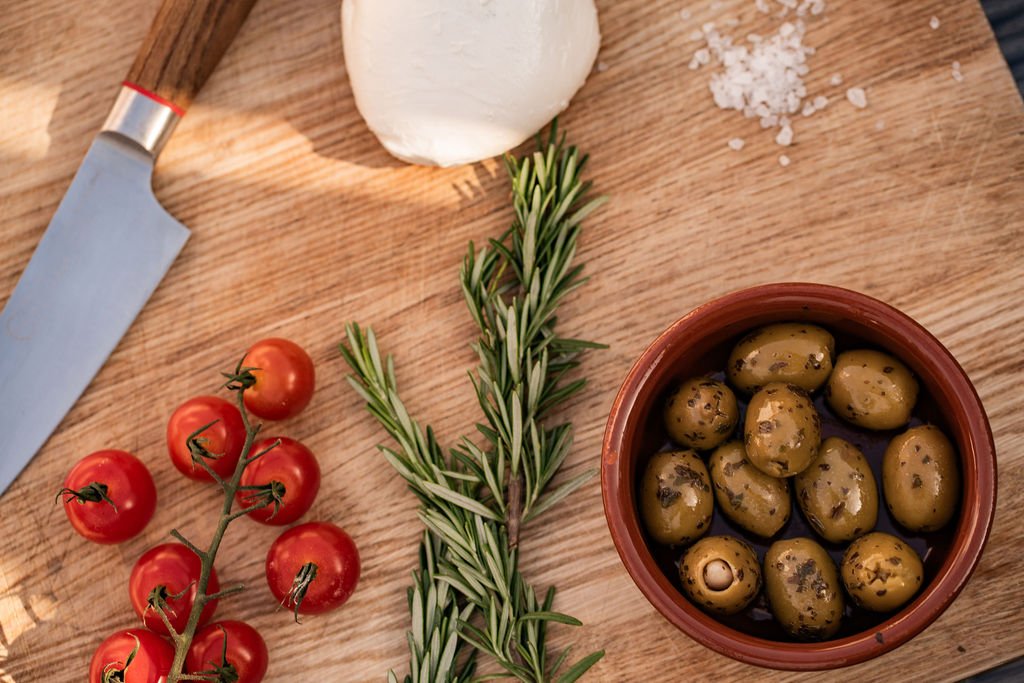 Cherry tomatoes on the vine, a sprig of rosemary, a ball of fresh mozzarella, and a bowl of green olives on a wooden cutting board.