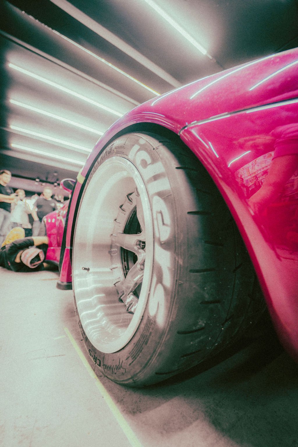Close-up of a pink Porsche RWB (Rubi) front tire and fender in a garage setting, with people in the background.