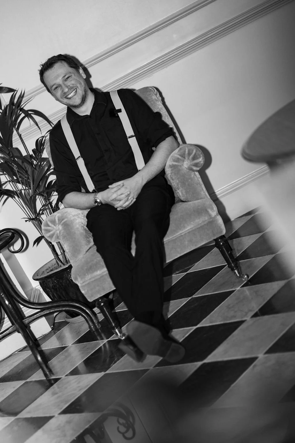 A smiling man sitting on a vintage armchair, wearing a black shirt with suspenders and black pants, in a room with checkered floor and decorative wall molding.