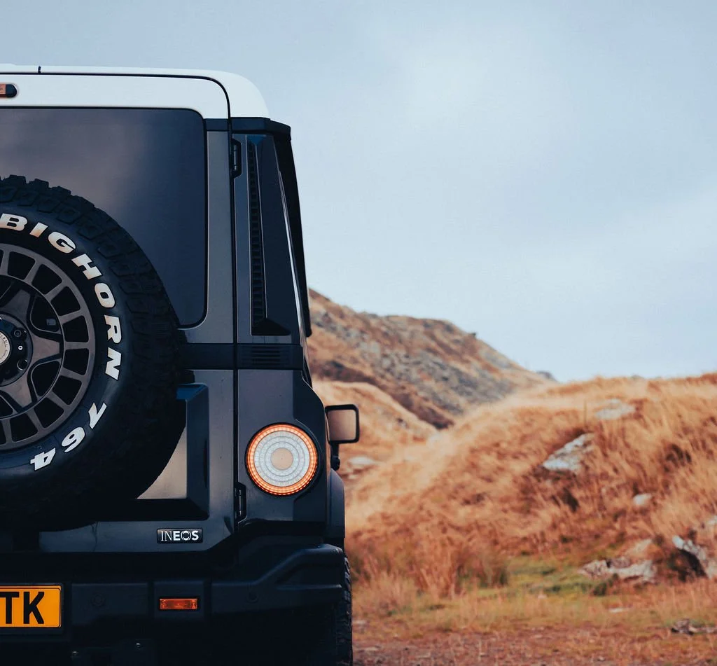Rear portion of a black INEOS Grenadier SUV with a spare tire mounted on the back, off-road terrain with rocks and dry grass in the background.