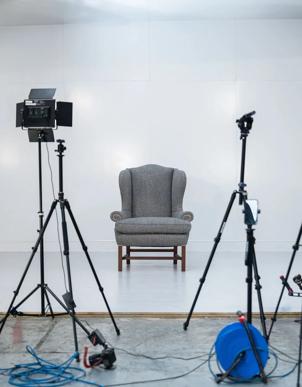An empty gray armchair centered in front of a plain white wall, with professional photography equipment like lights and tripods set up around it.