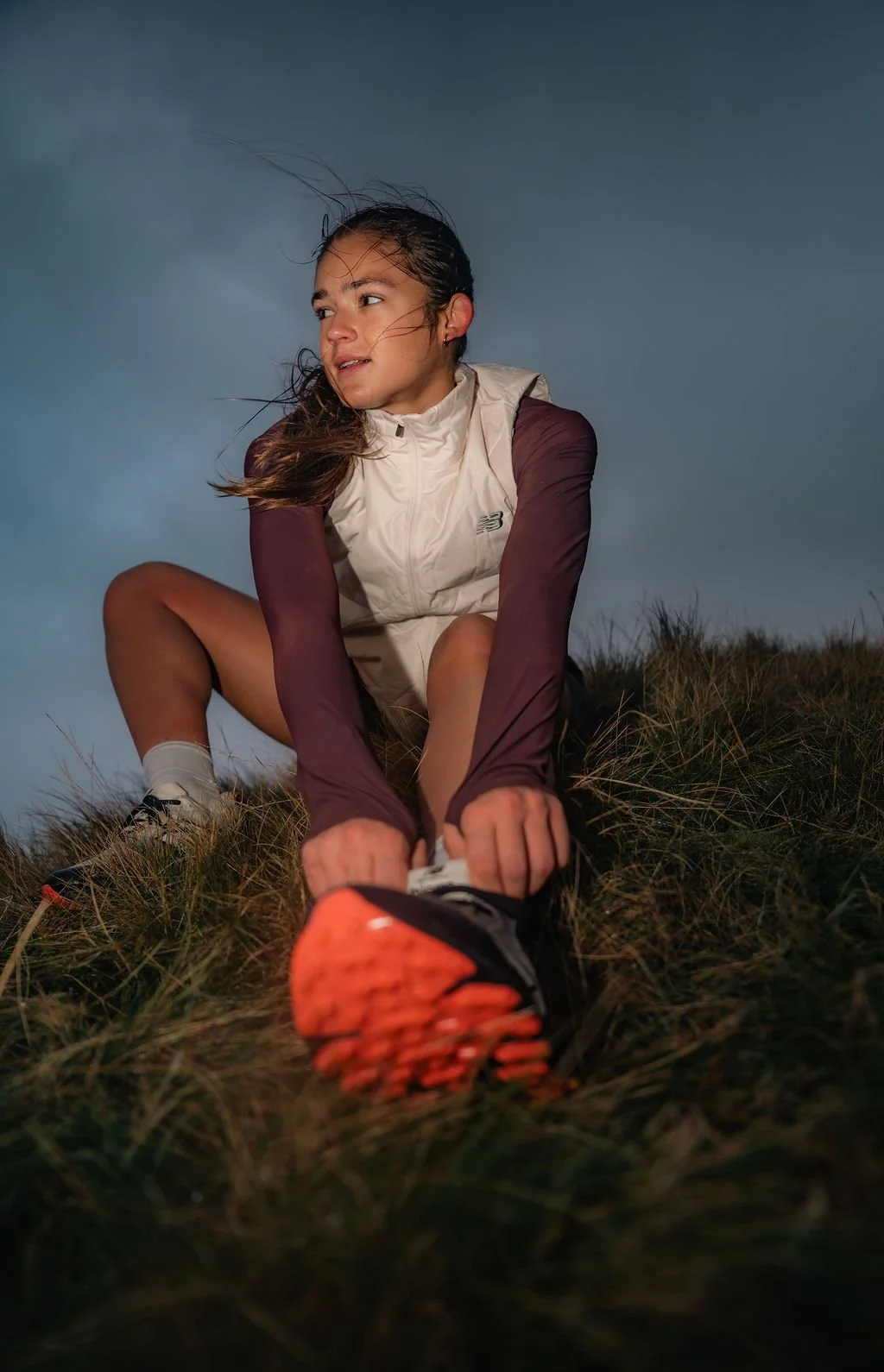 A young woman outdoors in a grassy field, tying her shoelace, under a cloudy sky, during dusk or dawn.