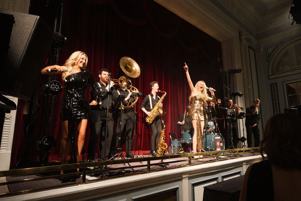A musical band performs on stage with a singer in a sequined gold dress singing into a microphone, in front of a red curtain.