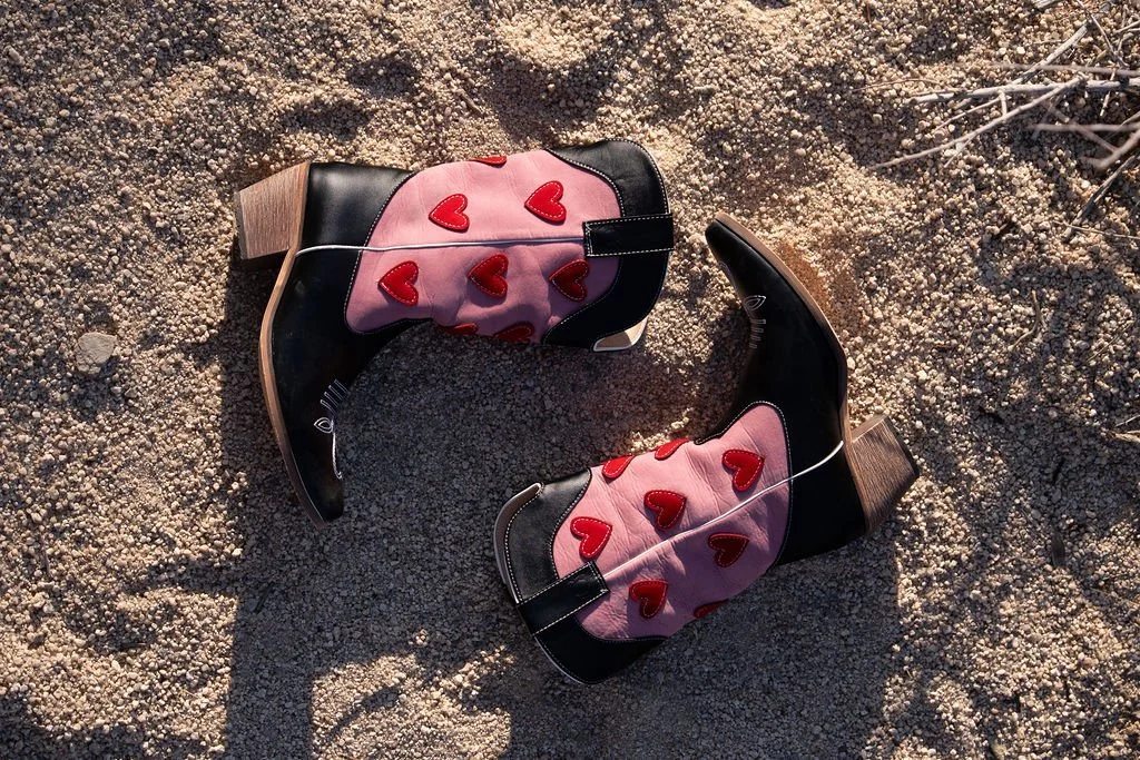 A pair of black cowboy boots with pink and red heart designs on the side, lying on sandy ground.