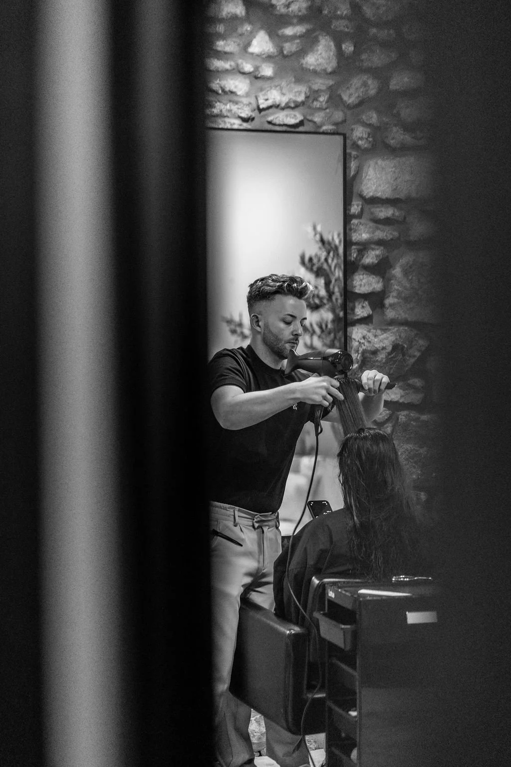 A hairstylist blow-drying a woman's hair in a salon with a stone wall background.