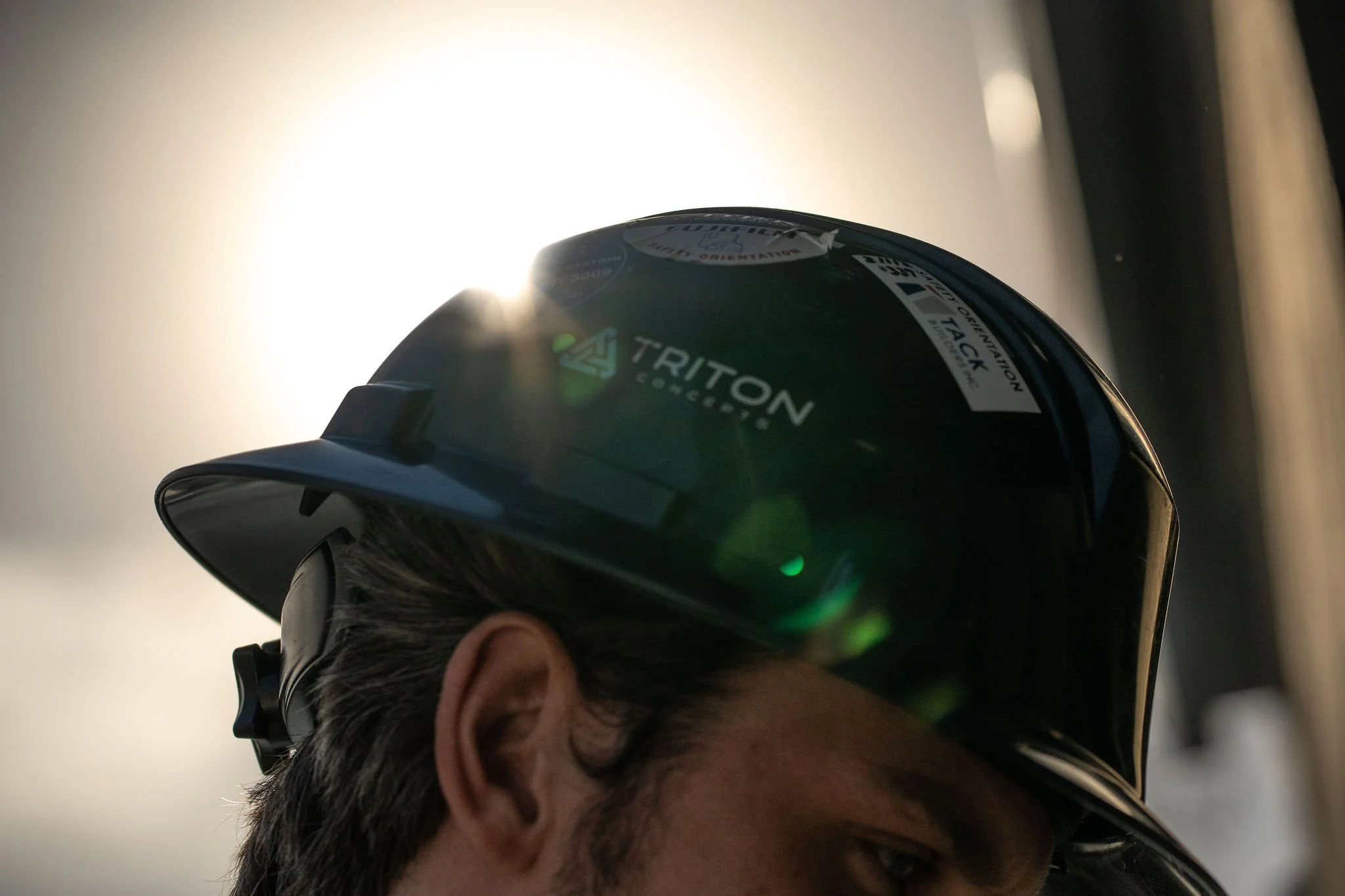 Close-up of a person wearing a black helmet with the Triton logo, taken from the side with sunlight behind.