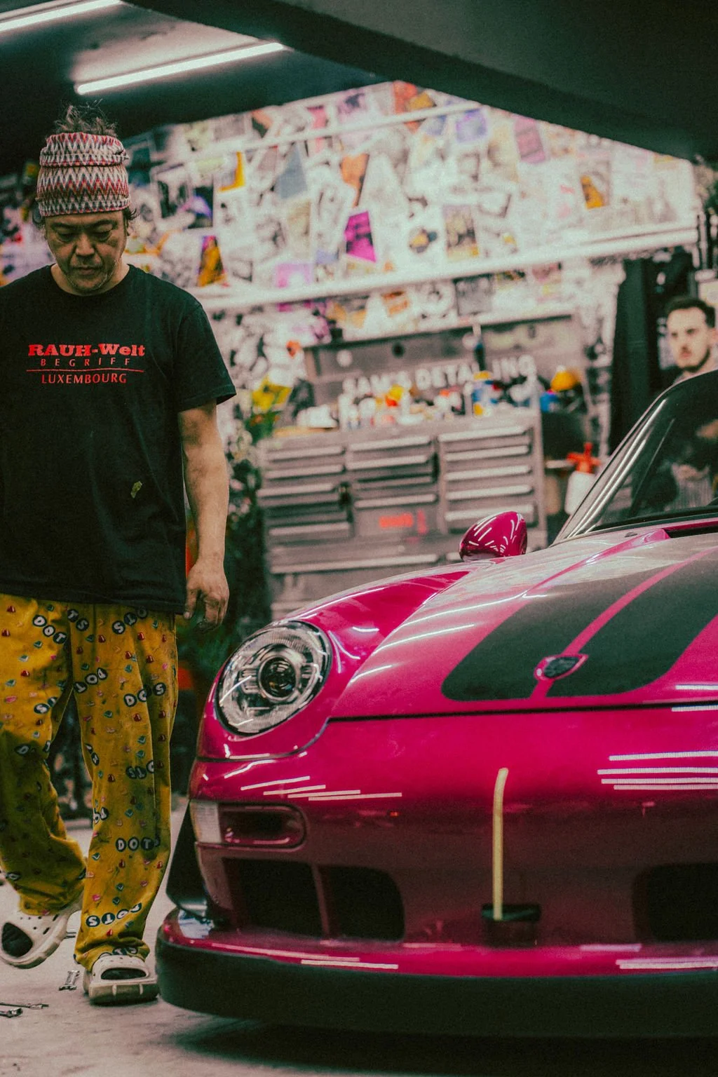 Nakaisan standing next to a pink RWB build in a garage or workshop, with a blurred person and tools and supplies in the background.