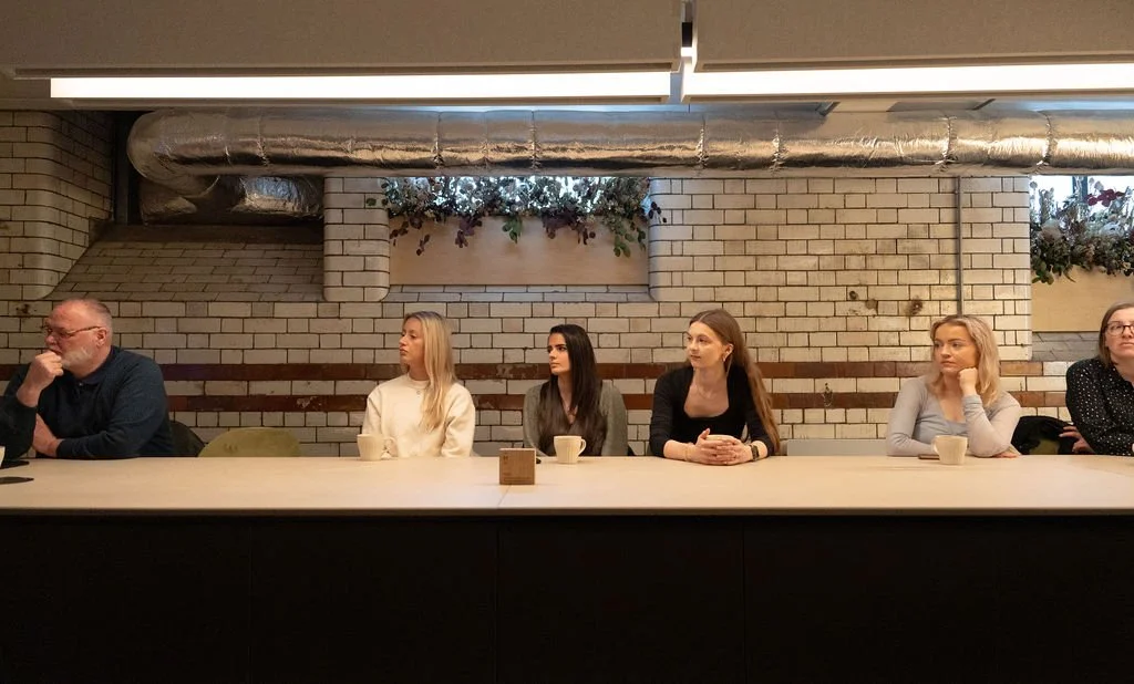 Six individuals sitting at a long table, with a brick wall and industrial ductwork in the background.