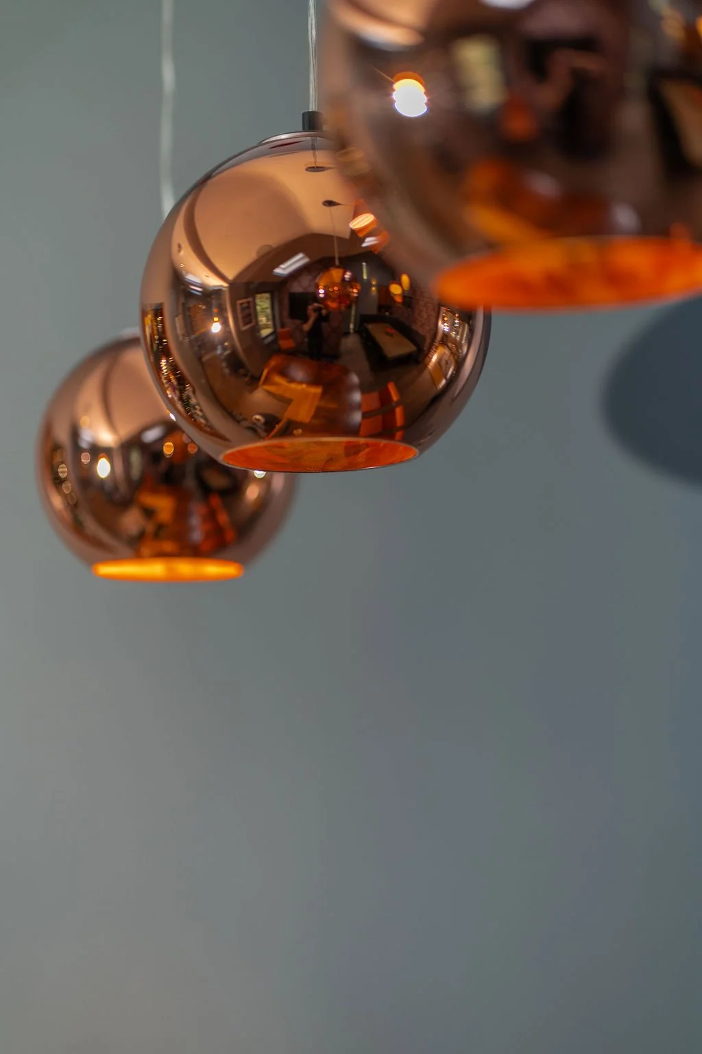 Close-up of three copper-colored spherical pendant lights hanging from the ceiling, reflecting the room's interior including a person taking the photograph, furniture, and windows.