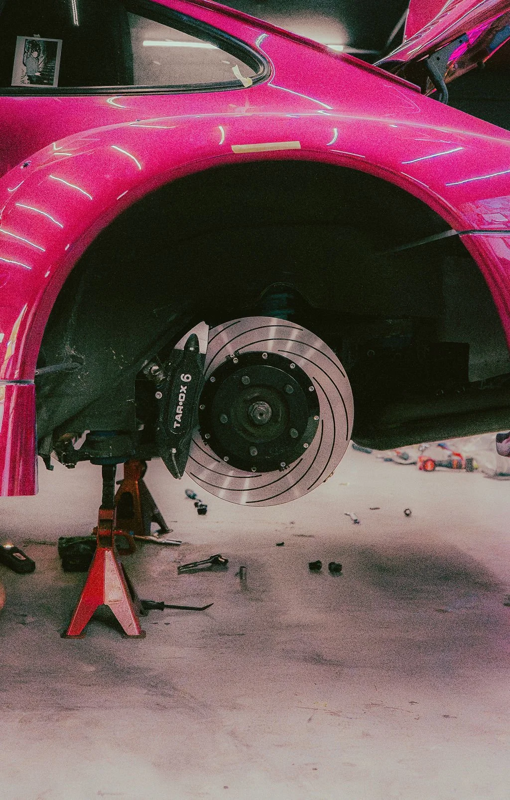 Close-up of a pink Porsche RWB with the front wheel removed, revealing a TAROX brake disc and caliper, supported by a red jack stand on a garage floor with tools scattered around.
