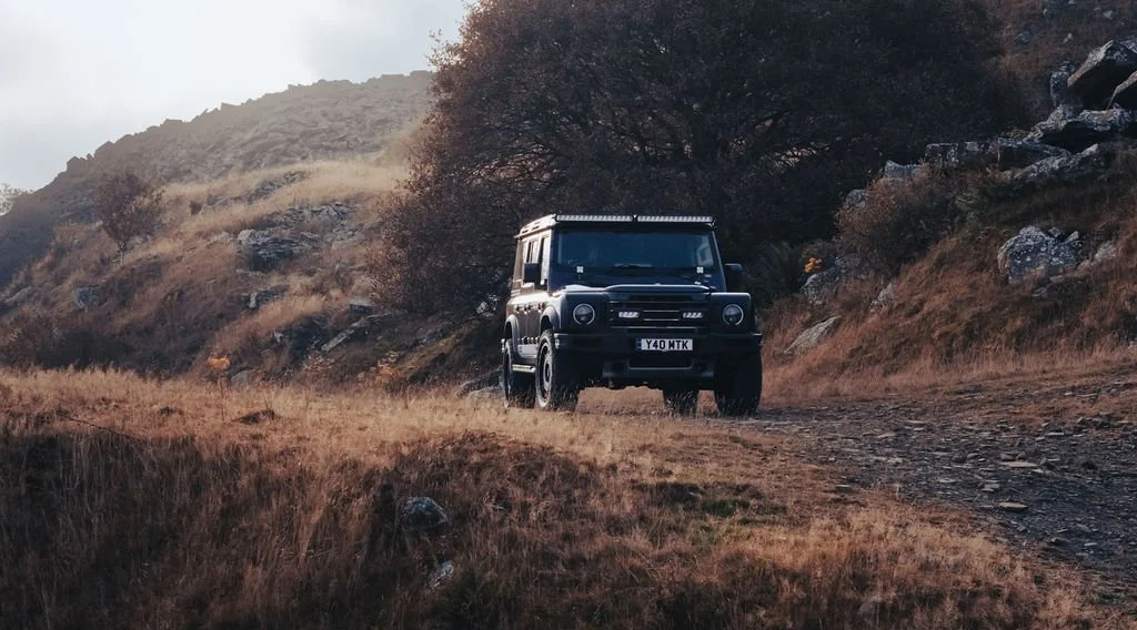 Black off-road vehicle driving on a dirt trail through a mountainous landscape with brown grass and sparse trees.
