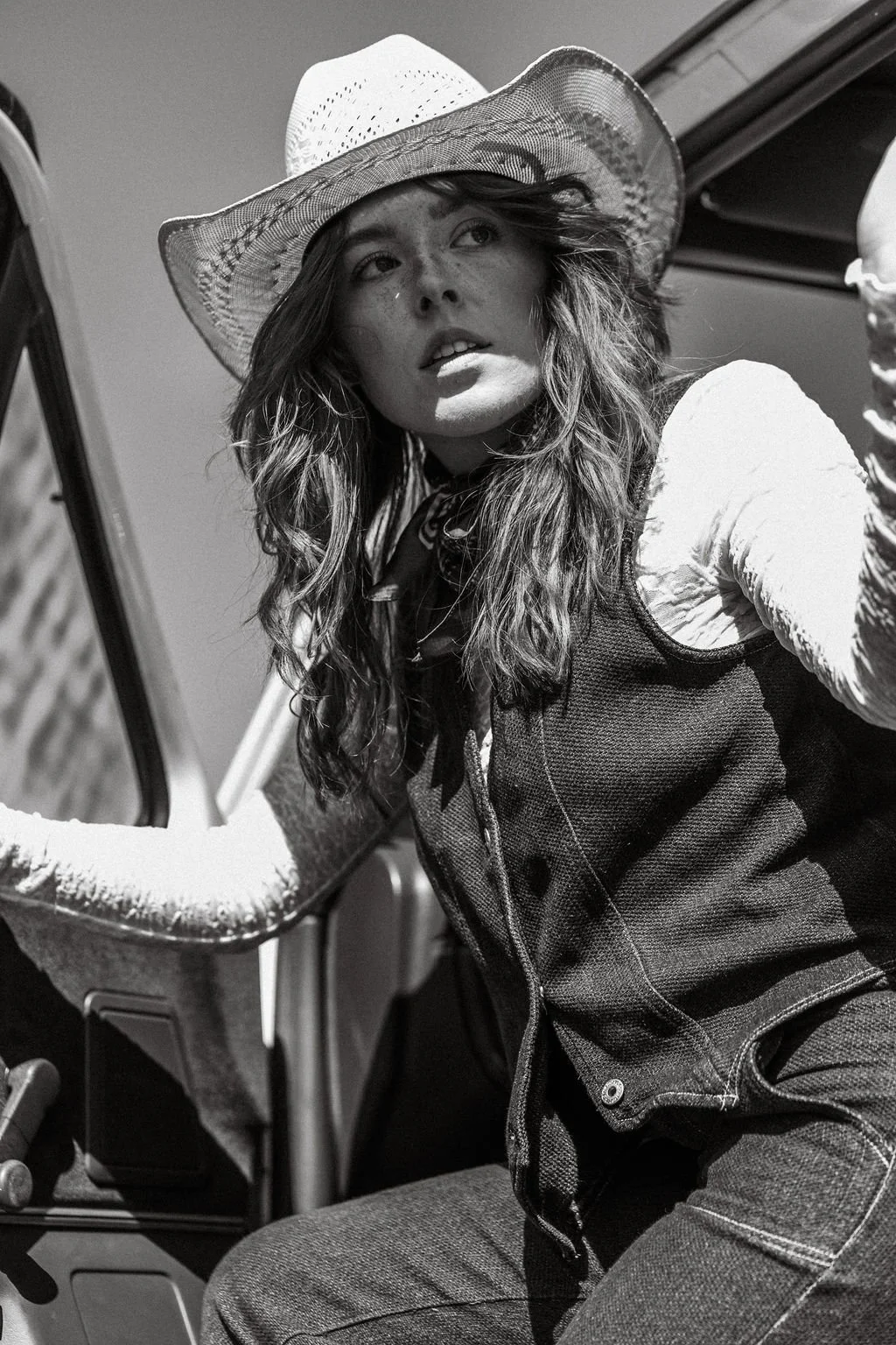 Black and white photo of a woman wearing a cowboy hat, a long sleeve shirt, and a vest, sitting in a vehicle and looking out of the window.