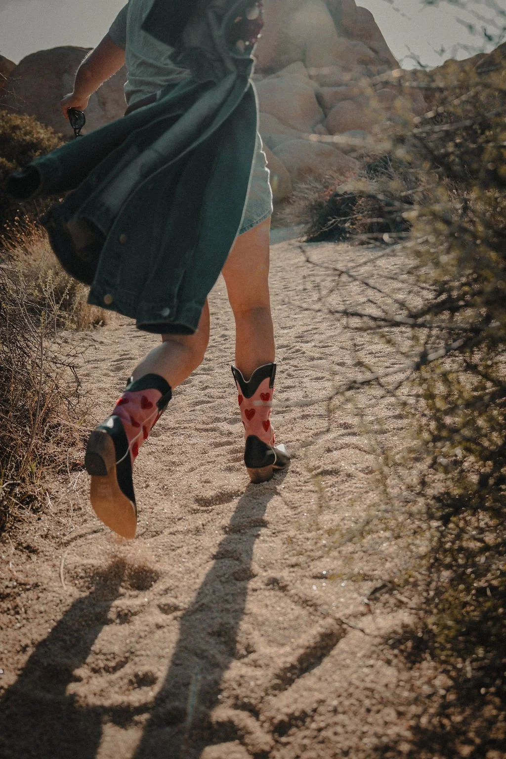 Person wearing cowboy boots with red hearts walking on a sandy trail in a desert landscape with rocks and bushes.