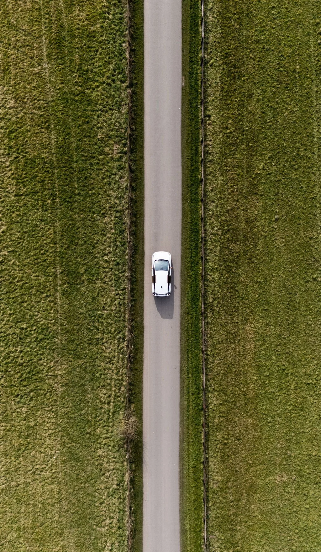 An aerial view of a white car driving down a narrow, paved country road bordered by green grassy fields.