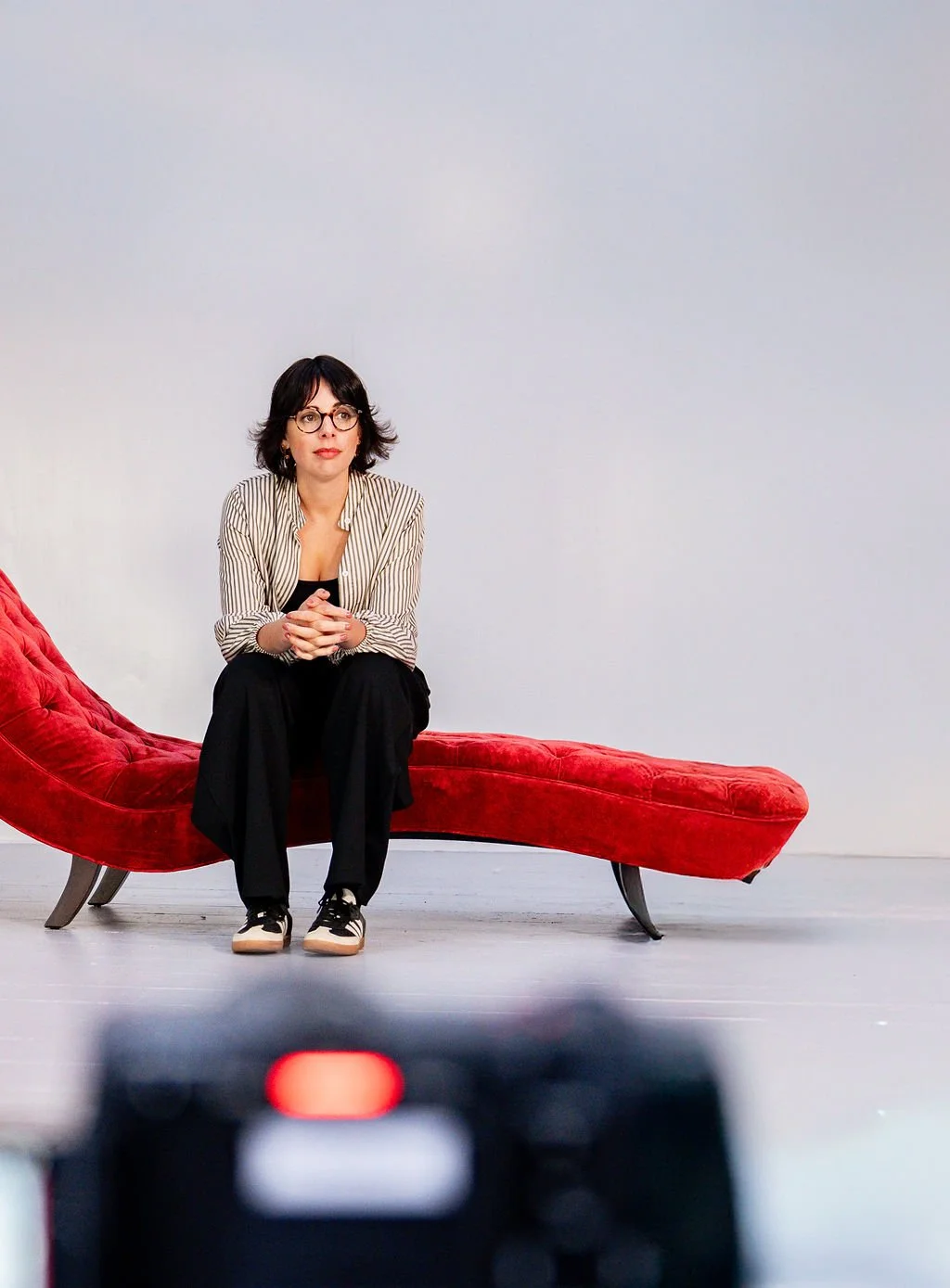 A woman with black hair and glasses sitting on a red velvet chaise longue with a plain white background.