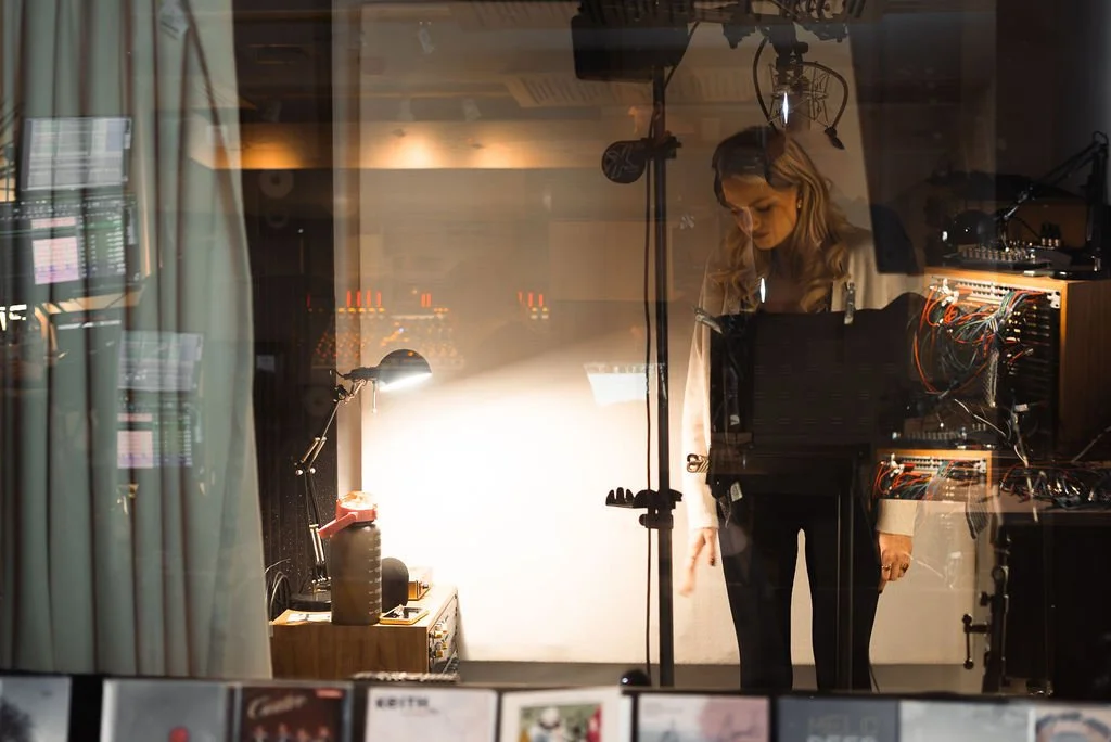 A woman inside a recording studio, wearing headphones, standing near a mixing console and audio equipment.