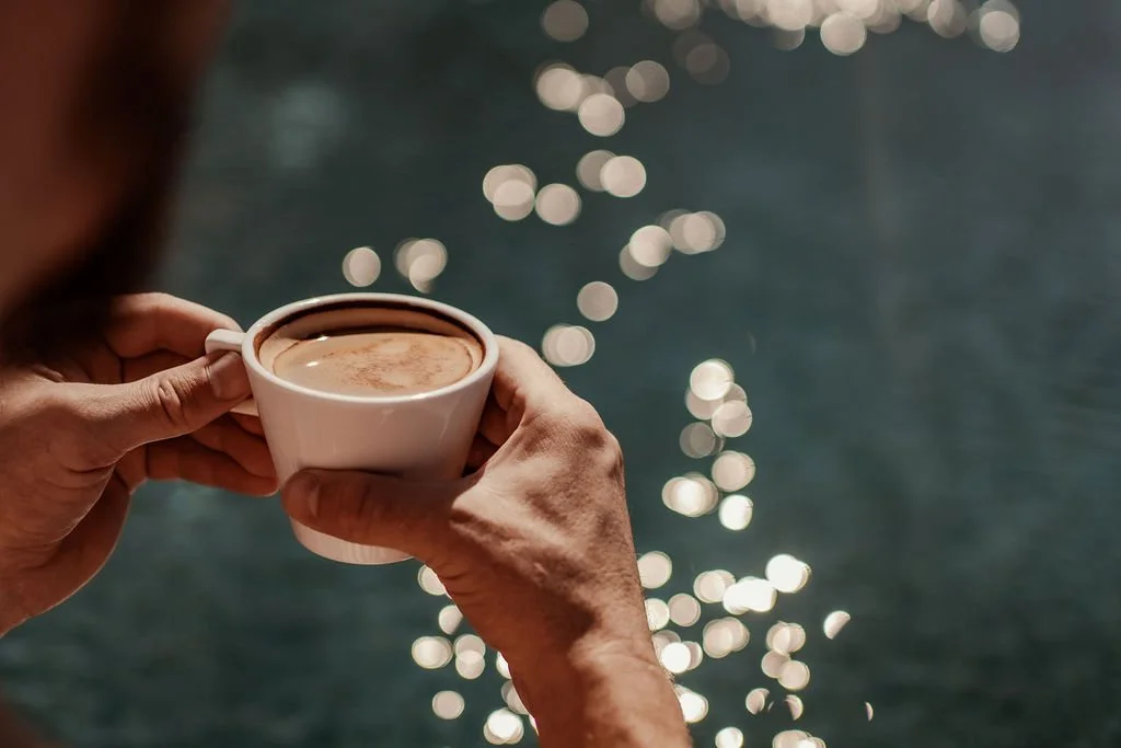 Person holding a cup of coffee near water with sunlight reflections