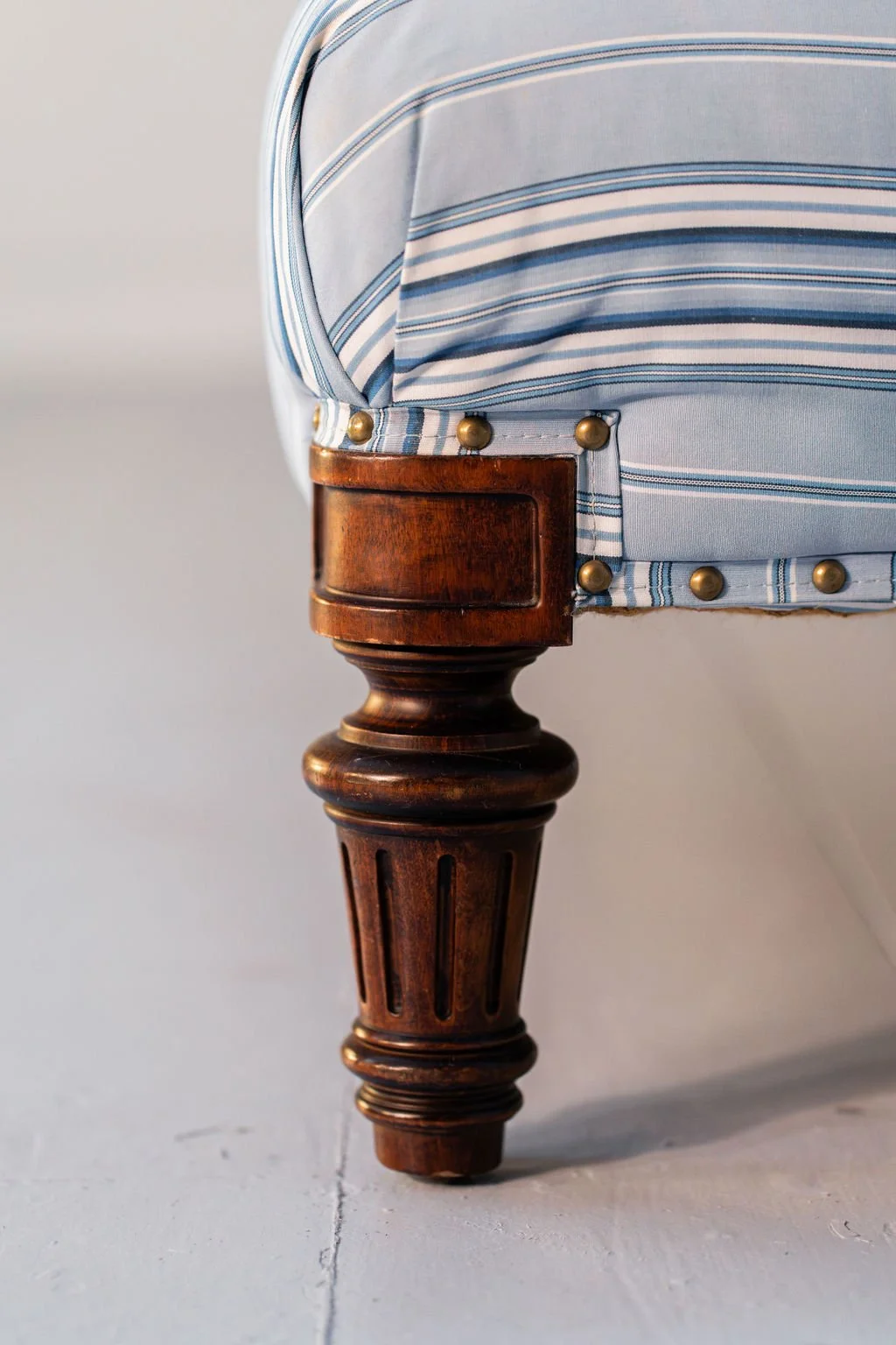Close-up of a wooden chair leg with a fabric trim in blue and white striped pattern, secured with brass studs.