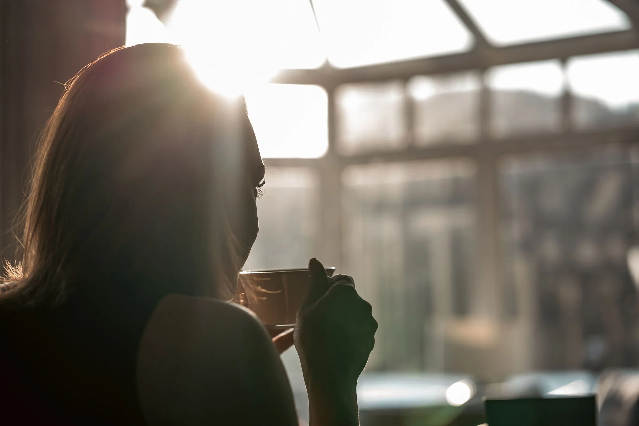 Silhouette of a woman with long hair drinking from a cup near a window with sunlight streaming in.