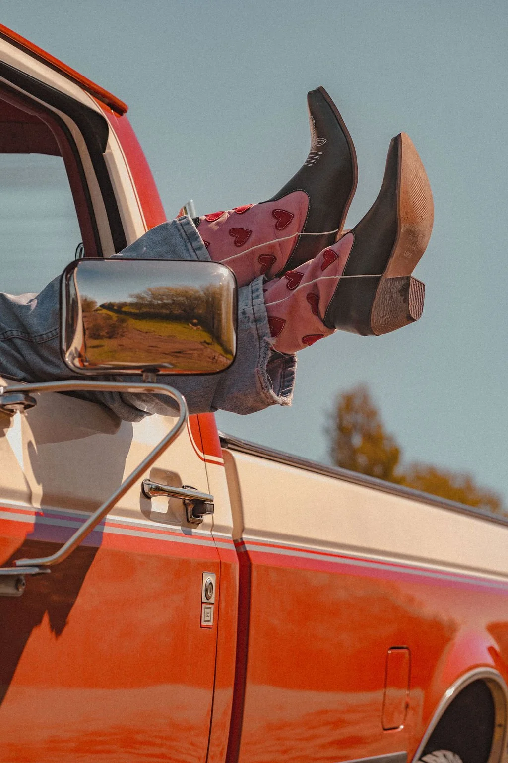 Close-up of a person with legs and feet sticking out of the window of a red vintage truck, wearing jeans, black cowboy boots with pink heart patterns. The side mirror reflects a landscape with trees and a dirt path.