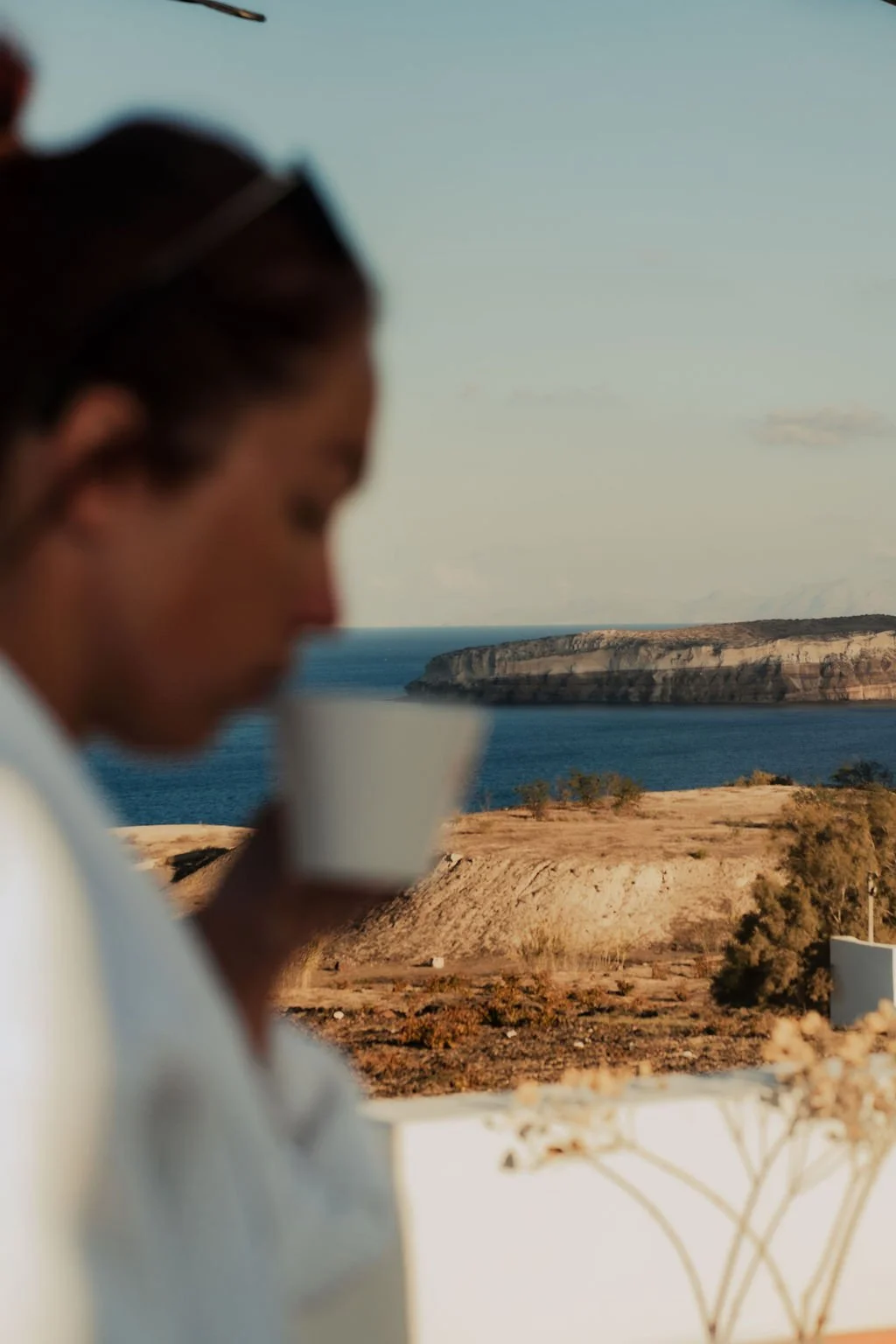 A person with dark hair wearing glasses is drinking from a white cup, with a scenic coastal landscape featuring cliffs, the sea, and a greek island in the background.