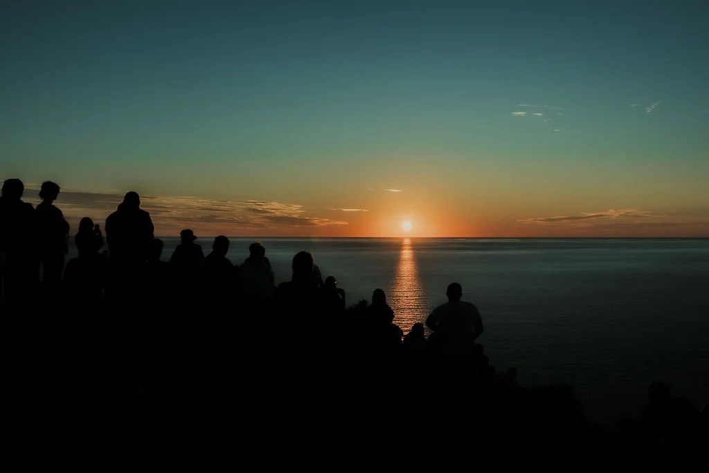 Silhouettes of people sitting and standing on rocks overlooking a sunset over the ocean, with the sun near the horizon and a reflection on the water.
