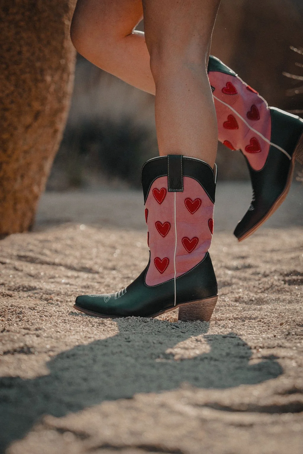 Close-up of a person's legs wearing cowboy boots with pink shaft and red hearts, standing on sandy ground near a rock.