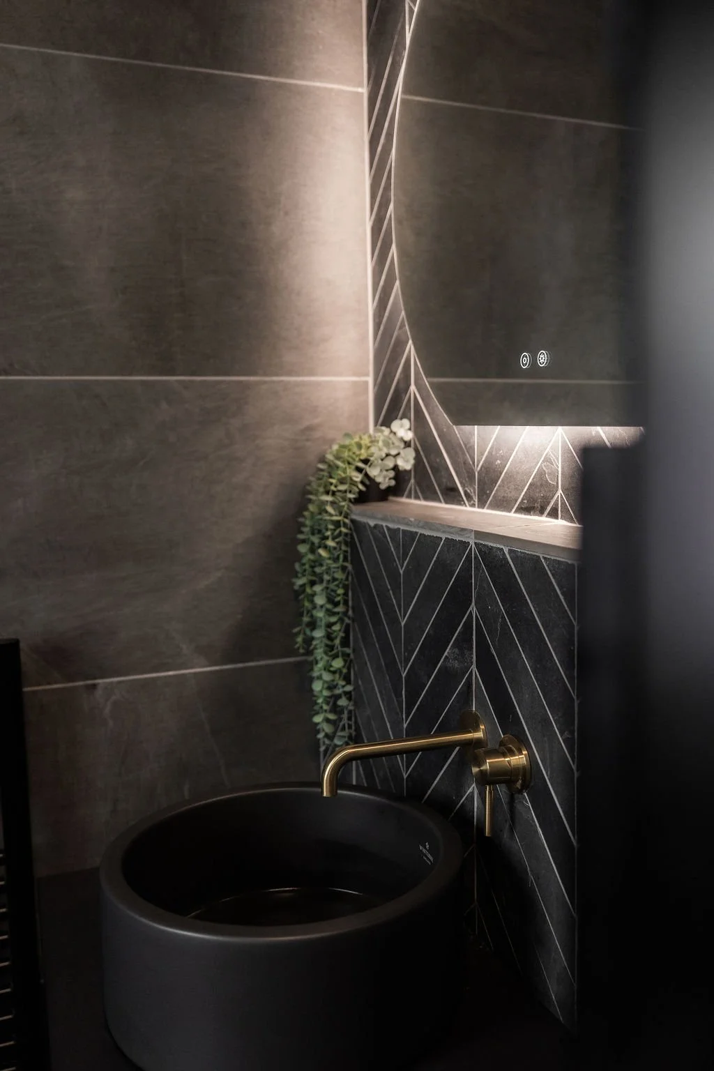 Part of a modern bathroom with dark tiled walls, a small black potted plant with trailing green foliage, and a black vessel sink with a gold faucet.