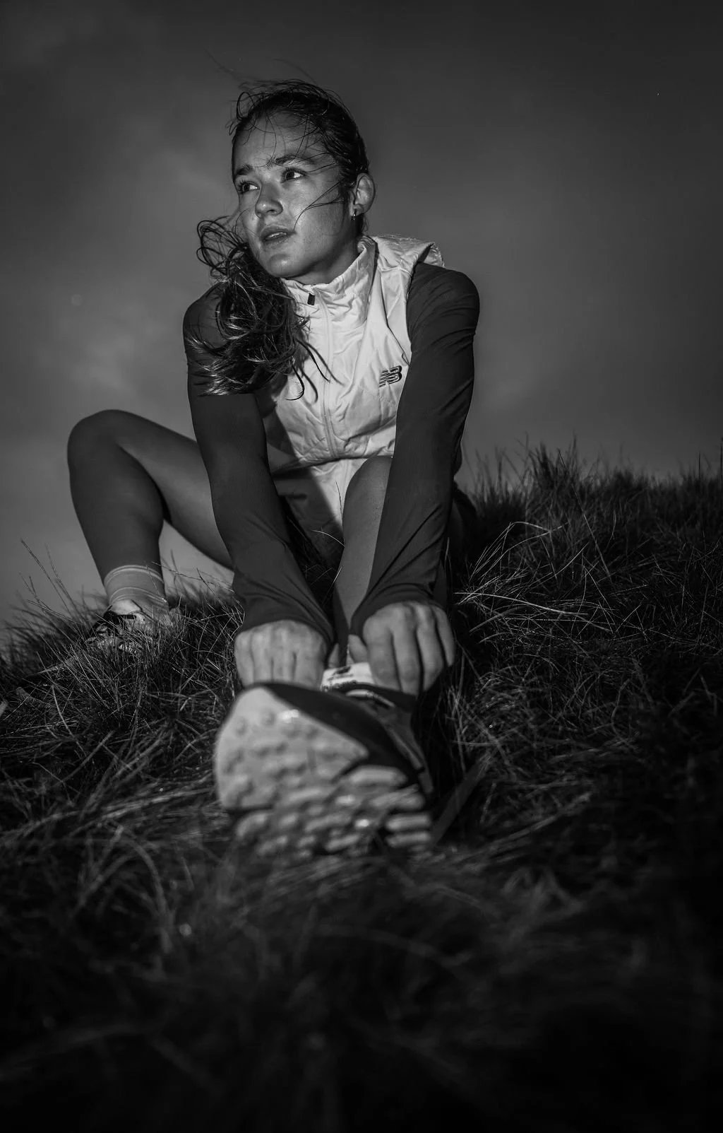 A woman outdoors, dressed in athletic wear, tying her shoelaces on a grassy hill during a cloudy day, black and white photograph.
