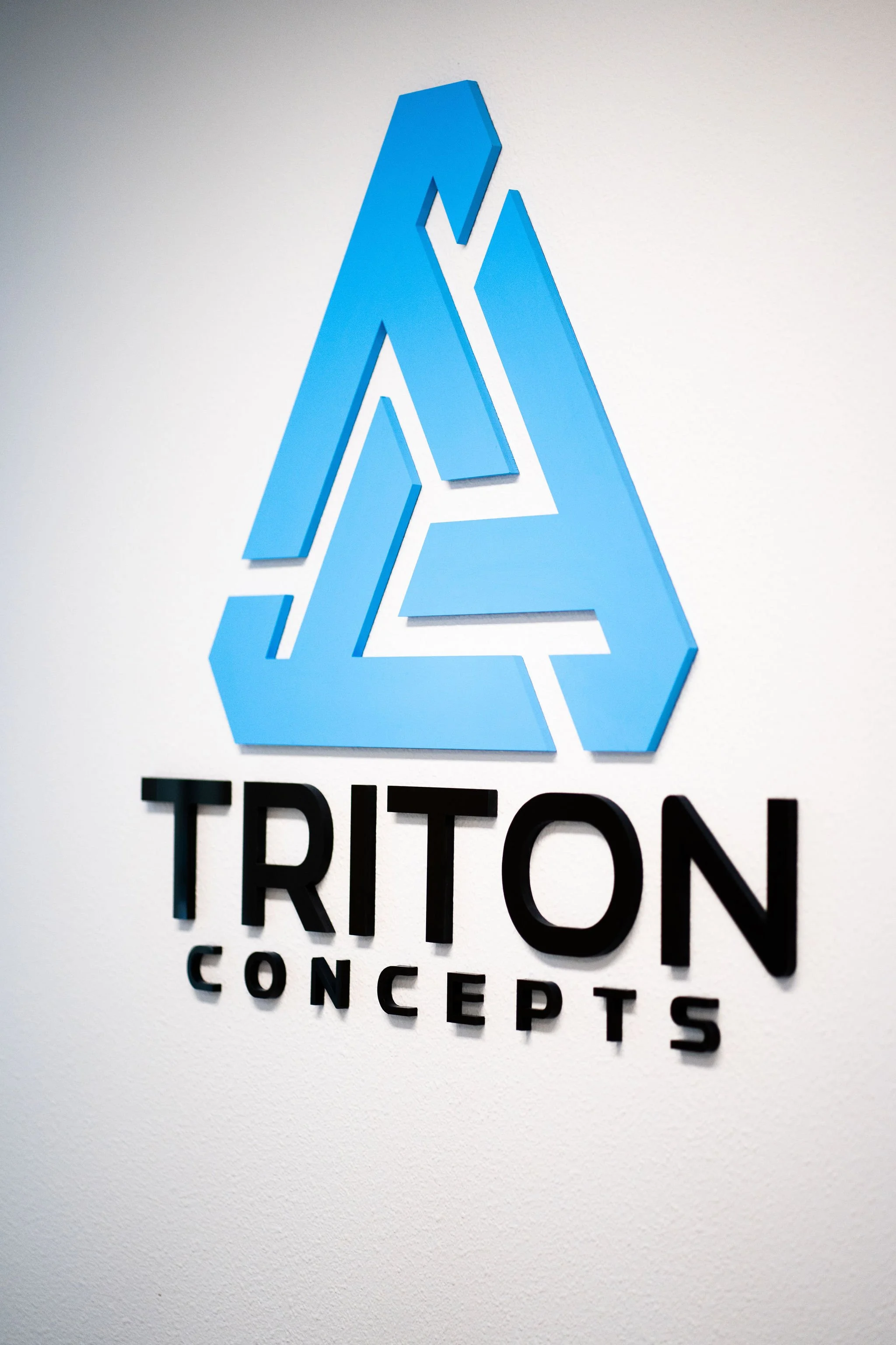 Blue logo of Triton Concepts on a white wall with the company name beneath it.